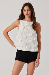 A young woman is wearing a sleeveless, white top decorated with 3D floral embellishments. The top has a round neckline and a loose fit. She pairs it with black shorts that have a textured fabric.